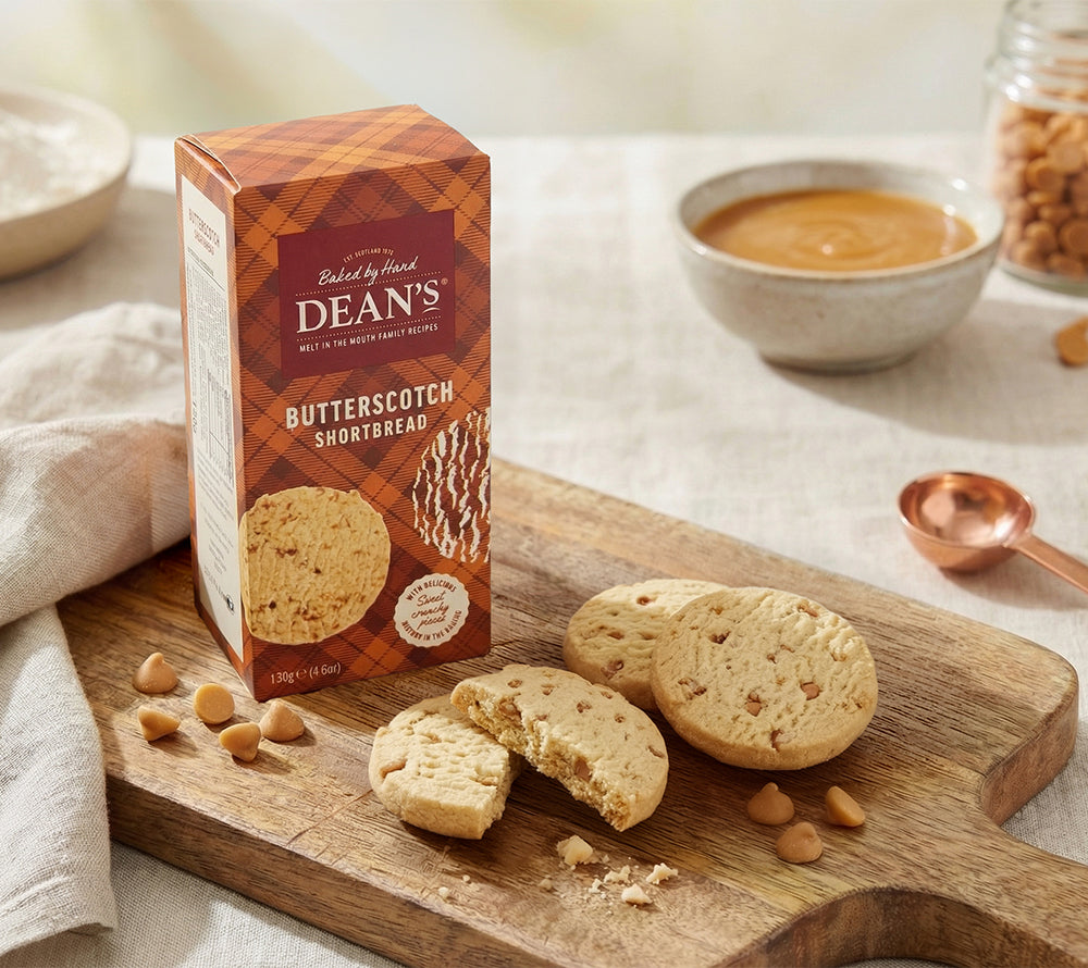 Gourmet‑Produktbild der Dean's Hand Baked Butterscotch Shortbread: Orange‑karierte Pappschachtel mit geöffneter Verpackung auf Schneidebrett, butterige Shortbread‑Kekse mit Butterscotch‑Chips, Schüssel mit warmer Karamellsauce, Löffel, weißes Tuch im Hintergrund