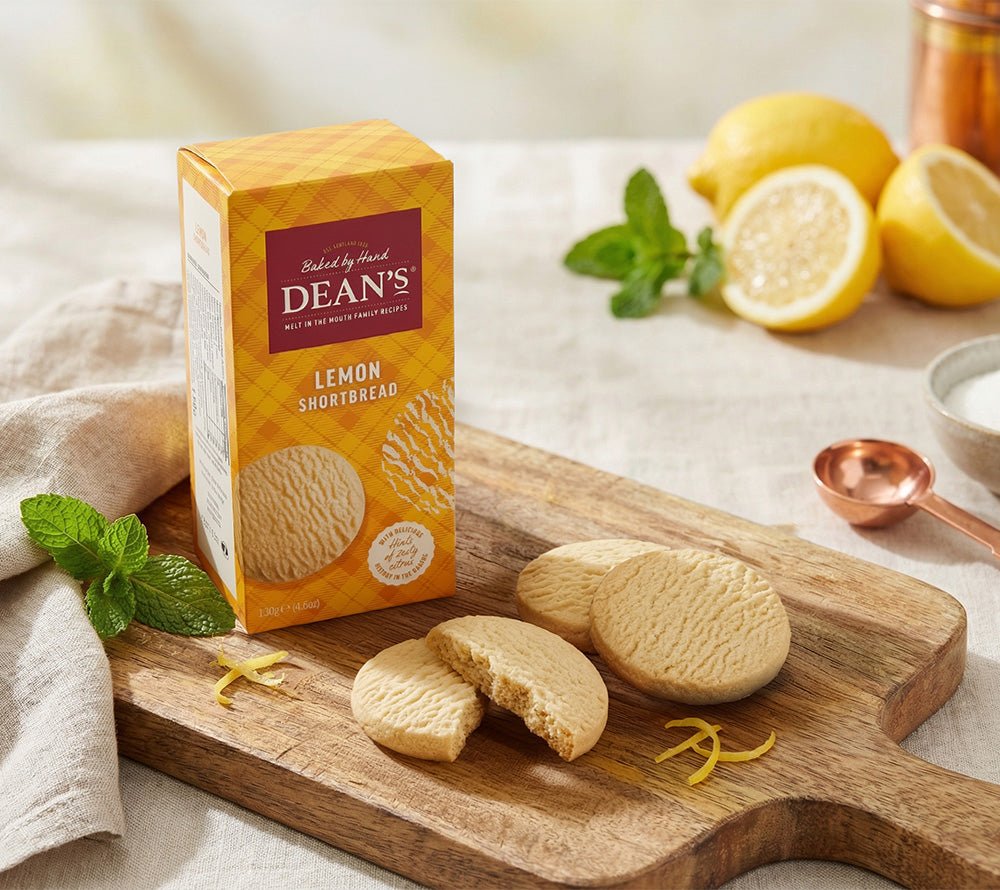 Dean's Hand Baked Lemon Shortbread in gelb‑karierten Papierschachtel auf weißem Tisch, neben Teekanne, Teetassen, Zitronenscheiben, Minzblättern und Blumen – idyllisches Tee‑Moment.